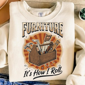 May include: A cream-coloured sweatshirt displaying a graphic of a wooden toolbox filled with carpentry tools. The text "FURNITURE" is above the toolbox, and "It's How I Roll" is below. The design has a vintage, rustic aesthetic.