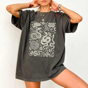 May include: A charcoal grey oversized t-shirt featuring a cream-coloured graphic print of multiple snakes intertwined with floral vines and leaves. The design covers the centre of the chest, showcasing various snake species in a detailed illustration.