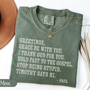 Olive green t-shirt with white text: "Greetings, Grace Be With You. I Thank God For You. Hold Fast To The Gospel. Stop Being Stupid. Timothy Says Hi. - Paul"