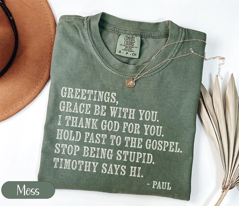 Olive green t-shirt with white text: "Greetings, Grace Be With You. I Thank God For You. Hold Fast To The Gospel. Stop Being Stupid. Timothy Says Hi. - Paul"
