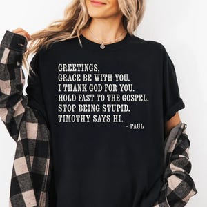 Black t-shirt with white text: "Greetings, Grace be with you. I thank God for you. Hold fast to the gospel. Stop being stupid. Timothy says hi. - Paul"