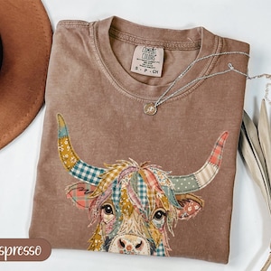 May include: Espresso-colored t-shirt with a colorful patchwork highland cow graphic. The shirt has a crew neck and short sleeves. A silver necklace with a small pendant rests on the shirt. The shirt is labeled "Comfort Colors" and is size S-CH.