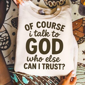 May include: A folded, cream-colored sweatshirt with the text "OF COURSE I talk to GOD who else CAN I TRUST?" in dark brown lettering. The sweatshirt is held by hands, and the background features a patterned fabric.