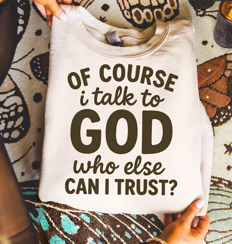 May include: A folded, cream-colored sweatshirt with the text "OF COURSE I talk to GOD who else CAN I TRUST?" in dark brown lettering. The sweatshirt is held by hands, and the background features a patterned fabric.