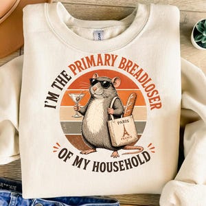 Op de afbeelding: Crèmekleurige sweatshirt met een afbeelding van een rat met een zonnebril, die een martini en een baguette vasthoudt. De rat draagt een draagtas met de tekst "Paris France". De tekst op de sweatshirt luidt "I'm the primary breadloser of my household."