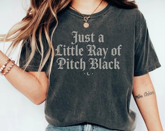 Puede incluir: Camiseta gris oscuro con la frase "Just a Little Ray of Pitch Black" en fuente gótica. La camiseta tiene un aspecto vintage y desgastado. El texto está centrado en el pecho.