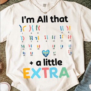 May include: White t-shirt with the text "I'm All that + a little EXTRA" in black, blue, orange, and green. The shirt features a colorful graphic of chromosomes, including a heart-shaped design with the number 21.