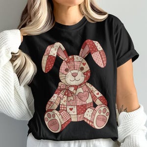 Op de afbeelding: Zwart T-shirt met een patchwork konijn ontwerp in rode en roze tinten. Het konijn heeft een strik, hart accenten en de woorden "Love", "Hugs" en "Kisses". Het ontwerp doet denken aan een knuffel.