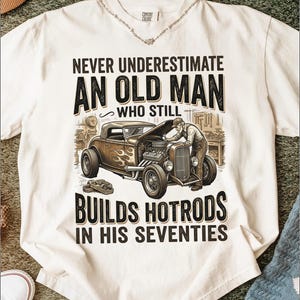 May include: White t-shirt with a graphic featuring a classic hot rod and a mechanic. The text reads "NEVER UNDERESTIMATE AN OLD MAN WHO STILL BUILDS HOTRODS IN HIS SEVENTIES."