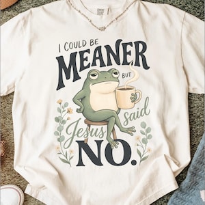 May include: An off-white short-sleeve t-shirt featuring a graphic of a green frog sitting on a stool, holding a steaming mug. The text reads I COULD BE MEANER BUT Jesus said NO. The design includes small floral sprigs on both sides of the frog.