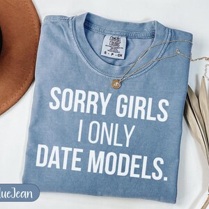 May include: A blue t-shirt with the text "SORRY GIRLS I ONLY DATE MODELS" in white block letters. A gold necklace with a pendant rests on the shirt. The shirt is folded and has a tag that says "COMFORT COLORS".