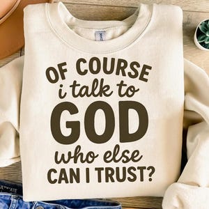 May include: Cream-colored sweatshirt with the text "OF COURSE i talk to GOD who else CAN I TRUST?" in dark brown lettering. The sweatshirt has a crew neck and long sleeves. A small potted plant is visible in the background.
