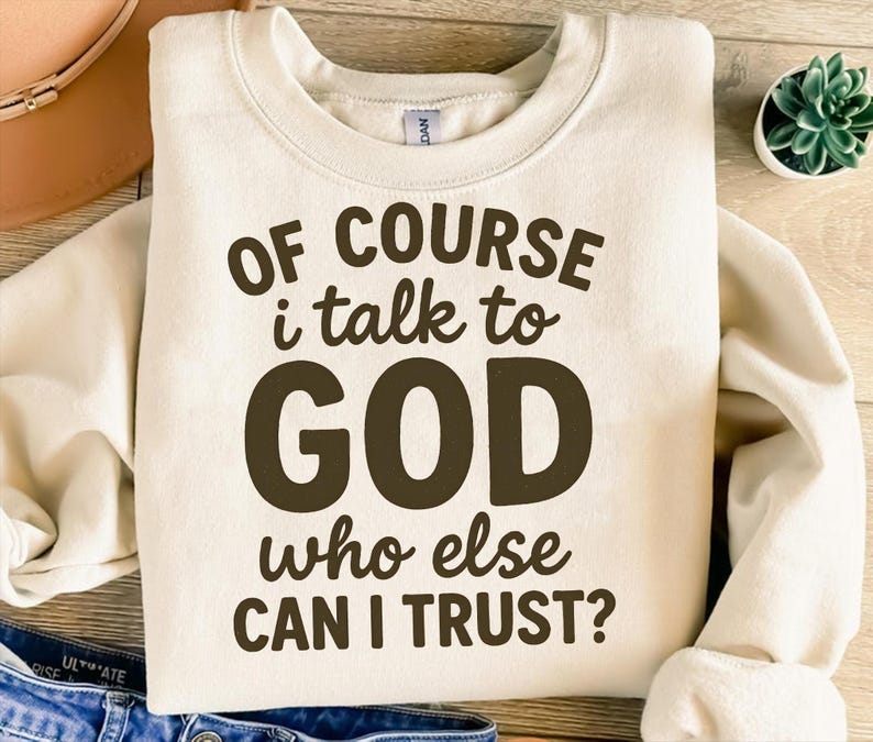 May include: Cream-colored sweatshirt with the text "OF COURSE i talk to GOD who else CAN I TRUST?" in dark brown lettering. The sweatshirt has a crew neck and long sleeves. A small potted plant is visible in the background.