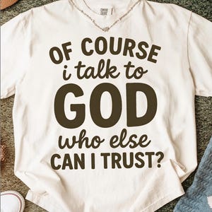 May include: Cream-colored t-shirt with the text "OF COURSE I talk to GOD who else CAN I TRUST?" in dark brown lettering. The shirt has a relaxed fit and a crew neck. A silver necklace is visible at the neckline.