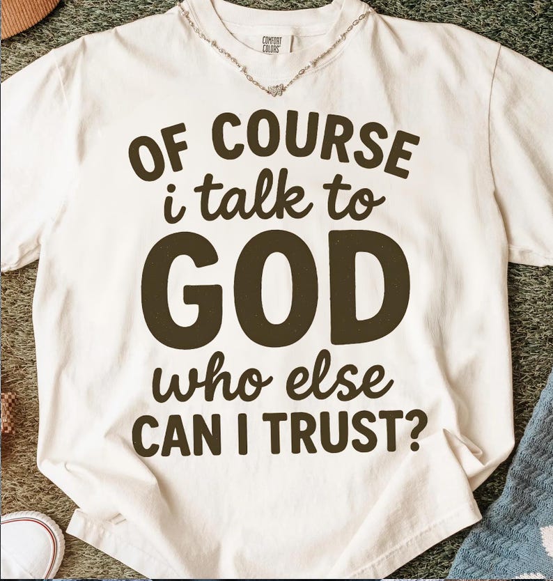 May include: Cream-colored t-shirt with the text "OF COURSE I talk to GOD who else CAN I TRUST?" in dark brown lettering. The shirt has a relaxed fit and a crew neck. A silver necklace is visible at the neckline.