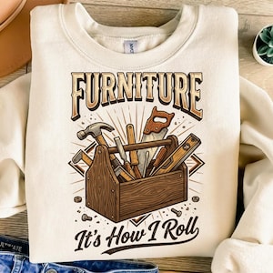 May include: A cream-coloured sweatshirt with a graphic of a wooden toolbox filled with tools, including a hammer and saw. The text "FURNITURE" is above the toolbox, and "It's How I Roll" is below the toolbox.