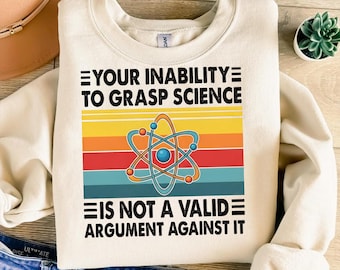 Puede incluir: Sudadera color crema con un gráfico colorido de un átomo y el texto "YOUR INABILITY TO GRASP SCIENCE IS NOT A VALID ARGUMENT AGAINST IT". El gráfico presenta un esquema de colores retro de amarillo, naranja, rojo y turquesa.