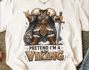 Puede incluir: Camiseta color crema con una ilustración de un vikingo. El vikingo está representado con armadura, sosteniendo una espada y un escudo, con el texto "PRETEND I'M A VIKING" debajo. El diseño incluye colores marrón, gris y beige.