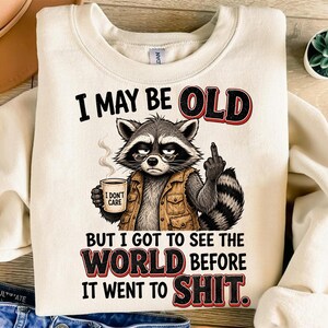 May include: A cream-coloured crewneck sweatshirt featuring a graphic of a raccoon wearing a tan vest and glasses. The raccoon holds a coffee mug labelled I DON'T CARE and gives the middle finger. Text reads: I MAY BE OLD BUT I GOT TO SEE THE WORLD BEFORE IT WENT TO SHIT.