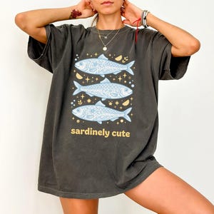 May include: Dark grey t-shirt with a design of three light blue fish, each with floral and scale patterns, and the text "sardinely cute" in yellow. The fish are set against stars and crescent moons.