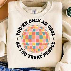 Puede incluir: Sudadera color crema con un gráfico de bola de discoteca colorida. El texto alrededor de la bola de discoteca dice "YOU'RE ONLY AS COOL AS YOU TREAT PEOPLE" en negro. El diseño incluye estrellas.