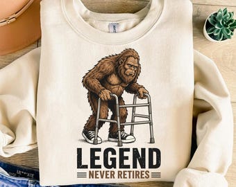 Puede incluir: Sudadera crema con un gráfico de Bigfoot usando un andador. El Bigfoot es marrón y lleva zapatillas negras. Debajo del gráfico, el texto dice "LEGEND NEVER RETIRES" en letras negras.