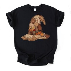 Puede incluir: Una camiseta negra de manga corta con un gráfico central de un sombrero de bruja hecho de varios parches de tela a cuadros en tonos marrón y beige. El diseño incluye pequeñas ilustraciones de calabazas naranjas y espigas de trigo.