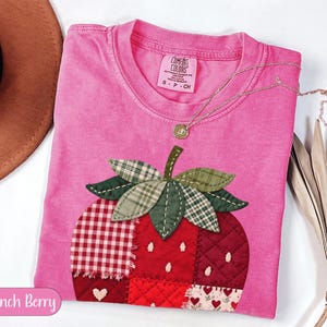 Könnte beinhalten: Ein leuchtend pinkfarbenes Kurzarm-T-Shirt mit einem großen, mittigen, erdbeerförmigen Aufnäher aus verschiedenen roten und weißen Stoffquadraten mit Karomustern, Steppungen und Herzmotiven. Das Shirt hat einen Rundhalsausschnitt und ein Comfort Colors-Etikett.