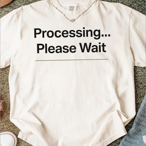 Puede incluir: Camiseta color crema con el texto negro "Processing... Please Wait" centrado. Cuello redondo y mangas cortas. Se aprecia un collar plateado. La tela parece suave.