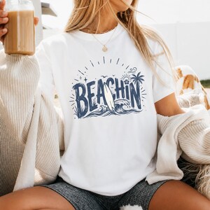 Może przedstawiać: Biała koszulka z granatową grafiką z napisem "BEACHIN" z deską surfingową, falami i palmą. Koszulka jest z krótkim rękawem i wykonana z miękkiego materiału.