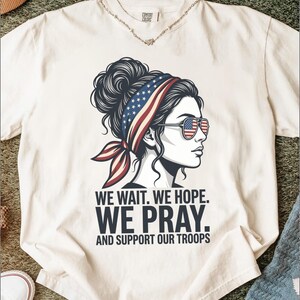Könnte beinhalten: Ein cremefarbenes Kurzarm-T-Shirt mit einer Grafik einer Person im Profil, die ein Stirnband und eine Sonnenbrille im US-Flaggen-Design trägt. Der Text lautet: WE WAIT. WE HOPE. WE PRAY. AND SUPPORT OUR TROOPS. Das Shirt hat einen Rundhalsausschnitt.