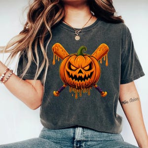 Puede incluir: Camiseta gris oscuro con un diseño temático de Halloween. El diseño incluye una calabaza naranja con una expresión amenazante, bates de béisbol cruzados y detalles naranjas goteando. Los bates tienen detalles morados y amarillos.
