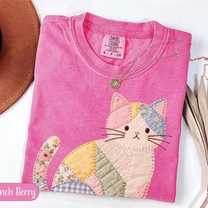Könnte beinhalten: Ein pinkfarbenes Comfort Colors T-Shirt mit einem Patchwork-Katzen-Design. Die Katze hat verschiedene Stoffmuster und Farben, darunter Pink, Blau, Gelb und Blumenmuster. Eine goldene Halskette mit Anhänger liegt über dem Shirt. Der Text "Crunch Berry" steht unten.