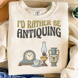 Puede incluir: Sudadera color crema con el texto "I'd Rather Be Antiquing" en verde oliva y negro. El diseño incluye ilustraciones de un reloj, una taza de té, una lámpara de aceite y un jarrón, todo en estilo vintage.