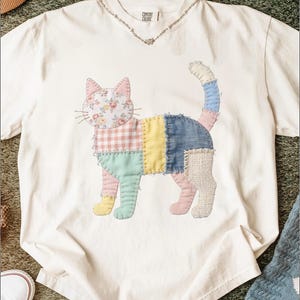 May include: Cream-colored t-shirt featuring a patchwork cat design. The cat is composed of various fabric patterns and colors, including floral, gingham, denim, and solid hues. The shirt has a V-neck and the text "COMFORT COLORS" is visible.