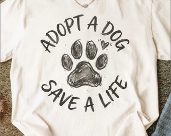 Puede incluir: Camiseta color crema con el texto negro "ADOPT A DOG SAVE A LIFE", junto con una huella de pata y un corazón. La camiseta está hecha de un material suave y es adecuada para uso casual.