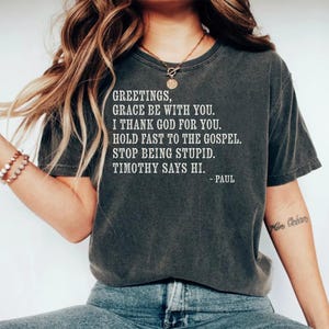 T-shirt with white text: "Greetings, grace be with you. I thank God for you. Hold fast to the gospel. Stop being stupid. Timothy says hi. - Paul".