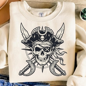 May include: A cream-colored crewneck sweatshirt featuring a large black graphic print of a skull wearing a pirate hat and an eye patch. Behind the skull is a compass rose design and two crossed cutlass swords, creating a detailed nautical illustration.