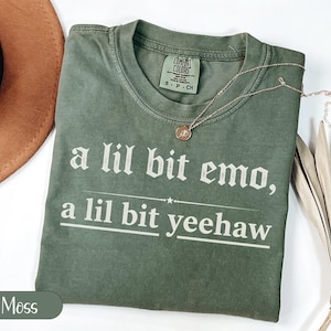 Może przedstawiać: Zielona koszulka w kolorze mchu z białym napisem "a lil bit emo, a lil bit yeehaw". Koszulka ma okrągły dekolt i krótkie rękawy. Złoty naszyjnik z wisiorkiem jest ułożony na koszulce.