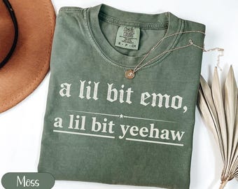 Puede incluir: Una camiseta verde musgo con el texto blanco "a lil bit emo, a lil bit yeehaw". La camiseta tiene cuello redondo y mangas cortas. Un collar dorado con un colgante está sobre la camiseta.