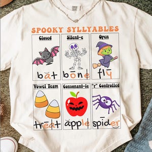 May include: Cream-colored t-shirt with a Halloween-themed graphic. The design features cartoon illustrations of a bat, skeleton, witch, candy corn, apple, and spider, with the words "SPOOKY SYLLABLES" and related vocabulary.