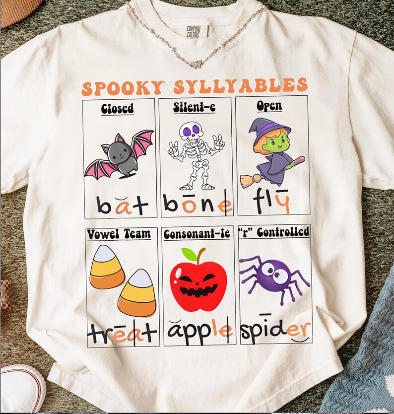 May include: Cream-colored t-shirt with a Halloween-themed graphic. The design features cartoon illustrations of a bat, skeleton, witch, candy corn, apple, and spider, with the words "SPOOKY SYLLABLES" and related vocabulary.