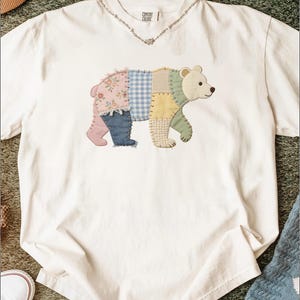 Könnte beinhalten: Weißes T-Shirt mit einem Patchwork-Bären-Design. Der Bär besteht aus verschiedenen Stoffmustern, darunter Blumenmuster, Jeansstoff und Vichy-Karo. Das Shirt hat einen Rundhalsausschnitt und kurze Ärmel. Der Text "COMFORT COLORS" ist auf dem Shirt aufgedruckt.