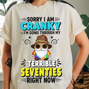 Könnte beinhalten: Cremefarbenes T-Shirt mit dem Text "SORRY I AM CRANKY - I'M GOING THROUGH MY TERRIBLE SEVENTIES RIGHT NOW." Das Design zeigt einen Cartoon-Zwerg mit weißem Bart, Sonnenbrille und einem Batik-Shirt.
