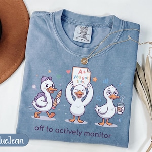 Puede incluir: Camiseta azul vaquero con un diseño de tres patos de dibujos animados. Un pato lleva un lápiz, otro sostiene un papel con "A+ you got this" y el tercero sostiene una taza de café. El texto "off to actively monitor" está debajo.