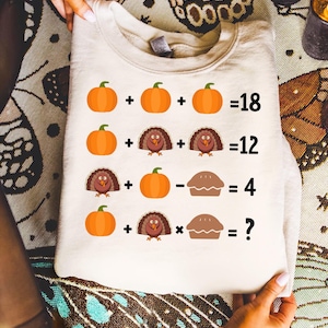May include: A cream-colored sweatshirt featuring a Thanksgiving-themed math problem with pumpkins, turkeys, and a pie. The equation includes the numbers 18, 12, 4, and a question mark. The sweatshirt is folded.