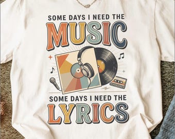 Puede incluir: Una camiseta color hueso con un estampado gráfico que incluye el texto "SOME DAYS I NEED THE MUSIC" y "SOME DAYS I NEED THE LYRICS". El diseño muestra un disco de vinilo, auriculares, un micrófono y un casete en colores retro.