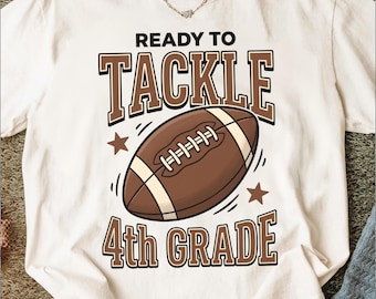 Puede incluir: Una camiseta de manga corta color blanco hueso con un gráfico marrón y crema de un balón de fútbol americano. El texto READY TO TACKLE 4th GRADE aparece en letras de bloque marrones. Dos estrellas marrones flanquean el gráfico del balón.