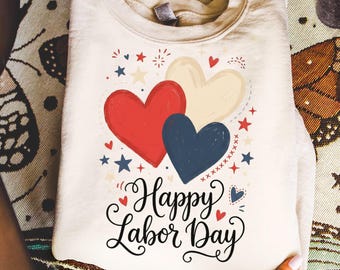 Puede incluir: Una sudadera de cuello redondo color crema con un estampado gráfico de tres corazones en rojo, azul marino y crema. El diseño incluye pequeñas estrellas y detalles de fuegos artificiales, con el texto Happy Labor Day en cursiva negra.