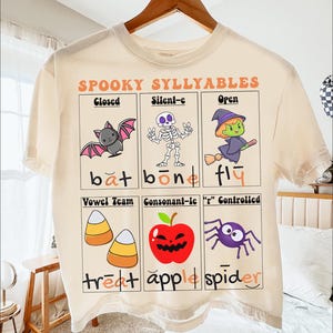 May include: Cream-colored t-shirt featuring a Halloween-themed graphic. The design includes illustrations of a bat, skeleton, witch, candy corn, apple, and spider, along with the text "SPOOKY SYLLYABLES". The shirt is hanging on a wooden hanger.